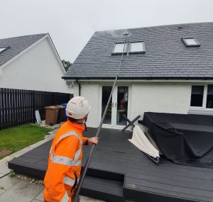Roof cleaner in high-visibility orange gear using a professional roof cleaning equipment on a grey slate roof with skylights on a single-storey home—demonstrating safe, ground-based roof cleaning.